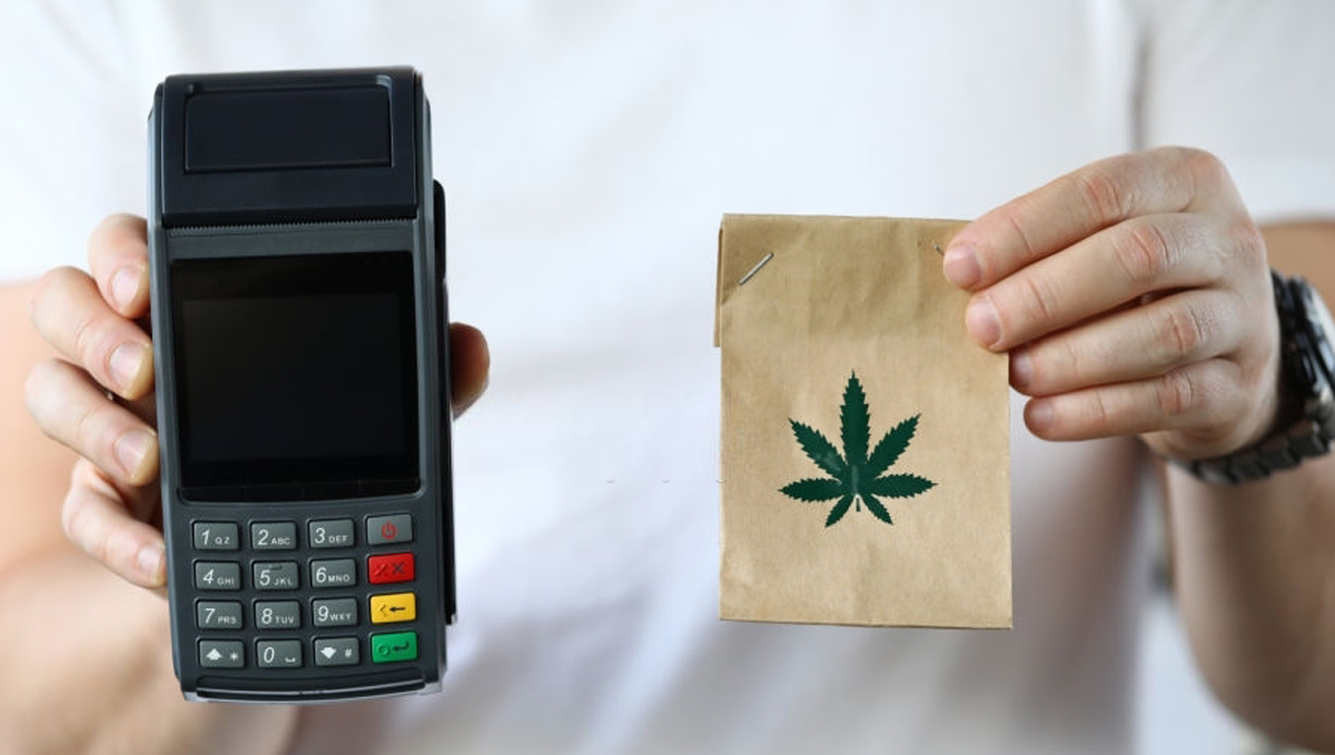 Visa Closes a Loophole That Allows Buying Weed With a Bank Card: A man holding a POS terminal in one hand and a brown paper pouch with a weed logo on it in the other Visa Closes a Loophole That Allows Buying Weed With a Bank Card: A man holding a POS terminal in one hand and a brown paper pouch with a weed logo on it in the other