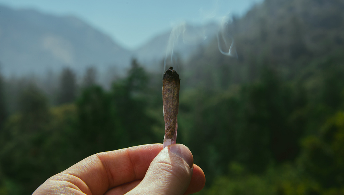 The Isle of Man Issues the First Medical Cannabis License: A closeup of a cannabis cigarette burning, with a mountainous landscape seen unfocused in the background