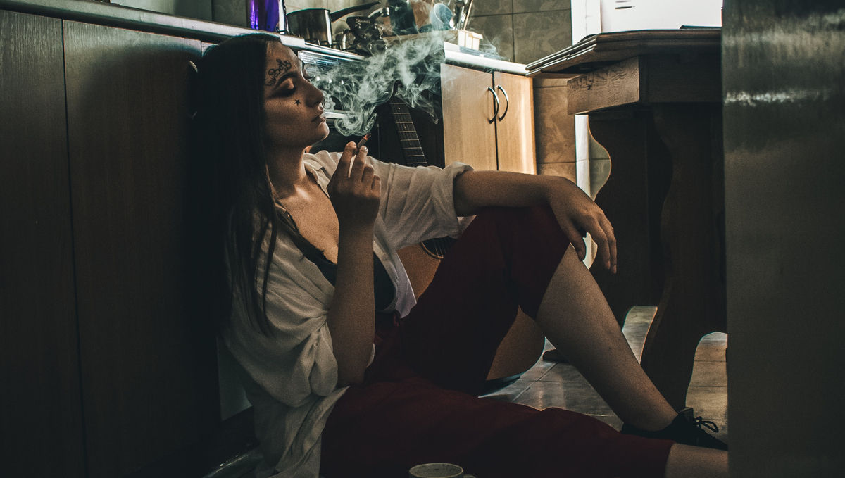 More Weed, Less Pills and Powders: Shifts in Drug Use During the Pandemic: A young despondent-looking woman sitting on the kitchen floor smoking a cigarette while her guitar stands idle right beside her