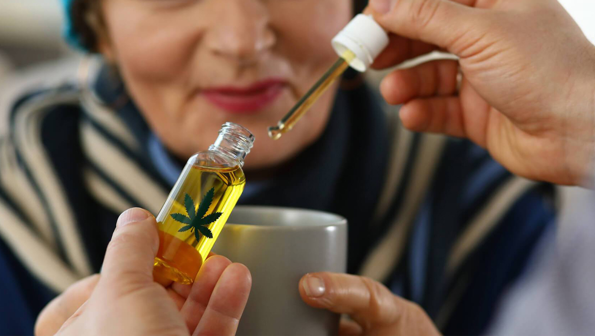 Cannabis Is Becoming More Popular Among the Elderly: A caregiver adding cannabis oil in a cup that an elderly woman holds
